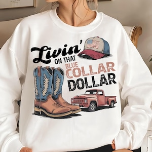 Peut inclure: Sweat-shirt blanc avec un motif graphique représentant des bottes de cowboy, une casquette de baseball avec un drapeau américain et un pick-up rouge. Le texte dit "Livin' on that Blue Collar Dollar."