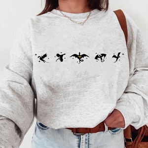 May include: A gray sweatshirt with a row of five black dragon silhouettes printed on the front. The dragons are flying in different directions, and the middle dragon has a yellow accent.