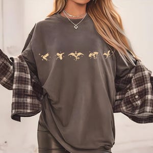 May include: A dark gray oversized t-shirt with a row of five gold dragon silhouettes printed on the front. The shirt is worn with a brown and white plaid flannel shirt tied around the waist.