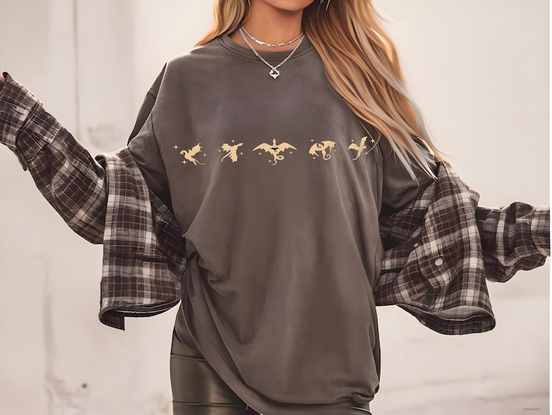 May include: A dark gray oversized t-shirt with a row of five gold dragon silhouettes printed on the front. The shirt is worn with a brown and white plaid flannel shirt tied around the waist.
