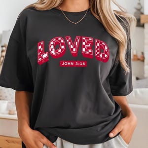 Peut inclure: T-shirt gris foncé avec le mot "LOVED" en rouge, souligné de blanc, avec un motif à carreaux à l'intérieur des lettres. En dessous, le texte "JOHN 3:16" en rouge. Le t-shirt a des manches courtes.