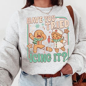 Könnte beinhalten: Hellgraues Sweatshirt mit weihnachtlichem Grafikdesign. Das Design zeigt zwei Lebkuchenmännchen, eines zerbrochen, das andere in Krankenschwesterntracht. Der Text lautet "Have You Tried Icing It?" im Retro-Stil. Enthält Sterne und einen grünen Rand.