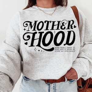 May include: Gray sweatshirt with the text "MOTHERHOOD" in black stylized lettering. The text "SOME DAYS I ROCK IT. SOME DAYS IT ROCKS ME. EITHER WAY WE'RE ROCKIN'" is printed below in smaller black lettering.