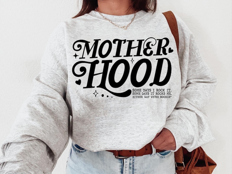 May include: Gray sweatshirt with the text "MOTHERHOOD" in black stylized lettering. The text "SOME DAYS I ROCK IT. SOME DAYS IT ROCKS ME. EITHER WAY WE'RE ROCKIN'" is printed below in smaller black lettering.