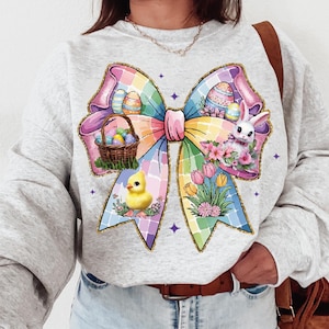 Peut inclure: Un sweat-shirt gris avec un motif de nœud d'Easter coloré. Le nœud est fait de tissu rose, bleu, jaune et vert et présente un panier d'œufs de Pâques, un poussin et un lapin. Le nœud est entouré de paillettes et d'étoiles.