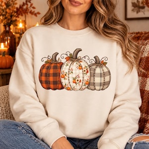Op de afbeelding: Een crèmekleurig sweatshirt met ronde hals en een afbeelding van drie pompoenen. De linkerpompoen heeft een oranje en zwart ruitpatroon, de middelste pompoen een witte bloemenprint en de rechterpompoen een grijs en wit ruitpatroon.