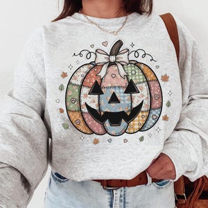 Peut inclure: Un sweat-shirt gris clair avec un motif de citrouille patchwork. La citrouille a un visage souriant et est décorée de motifs floraux, à carreaux et en forme de cœur. Le motif comprend un nœud, des feuilles et des étoiles, parfait pour l'automne.