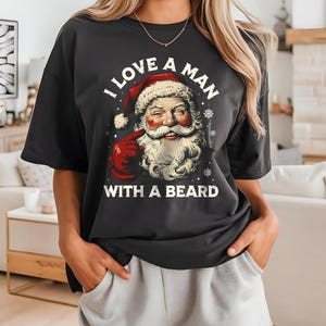 Puede incluir: Camiseta negra con un gráfico de Papá Noel, con barba blanca, gorro y guantes rojos. El texto "I LOVE A MAN WITH A BEARD" está impreso encima y debajo de la ilustración de Papá Noel en blanco.