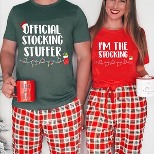 May include: Matching Christmas pajamas set. One person wears a green t-shirt with "OFFICIAL STOCKING STUFFER" text, and the other wears a red t-shirt with "I'M THE STOCKING" text. Both wear red and white plaid pants. Each holds a mug.