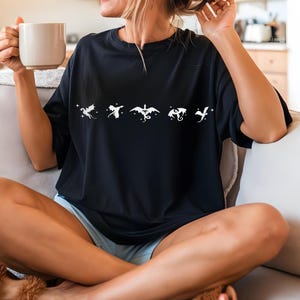 May include: A black t-shirt with a white graphic of five dragons in a row. The dragons are stylized and have a whimsical feel. The person wearing the shirt is sitting on a white couch.