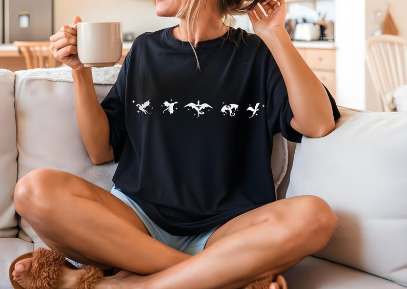 May include: A black t-shirt with a white graphic of five dragons in a row. The dragons are stylized and have a whimsical feel. The person wearing the shirt is sitting on a white couch.