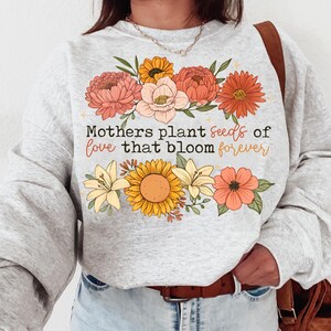 Peut inclure: Un sweat-shirt gris avec un motif floral et le texte "Mothers plant seeds of love that bloom forever".