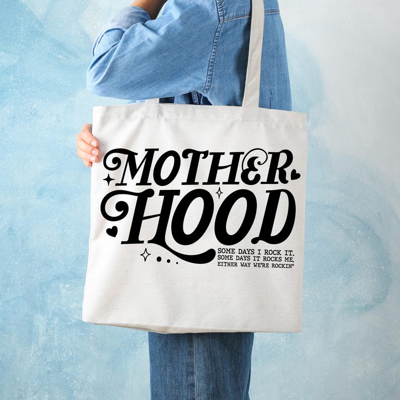 May include: White canvas tote bag with black text that reads "MOTHERHOOD" in a retro font. Below the text is a smaller font that reads "SOME DAYS I ROCK IT. SOME DAYS IT ROCKS ME. EITHER WAY WE'RE ROCKIN'"