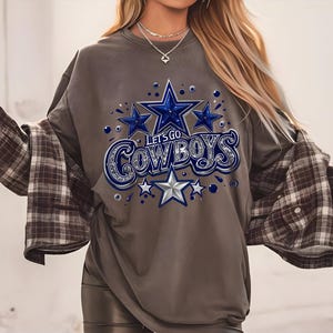 以下が含まれることがあります： グレーのトレーナーに、星と「Let's Go Cowboys」の文字が青と銀で描かれたグラフィックがプリントされています。
