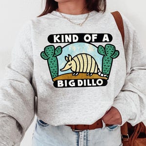 May include: A light gray sweatshirt with a graphic featuring an armadillo, cacti, and the text "KIND OF A BIG DILLO." The armadillo is tan and yellow, set against a blue sky and brown ground. Two green cacti flank the armadillo.