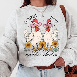 Peut inclure: Un sweat-shirt gris avec un motif de deux poulets portant des lunettes en forme de cœur. Le texte sur le sweat-shirt est "Oops! I got another chicken."