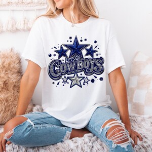 以下が含まれることがあります： 白いTシャツに、青と銀色のグラフィックで「Let's Go Cowboys」と星が描かれています。Tシャツは、白と茶色のふわふわした表面に座っている人が着ています。
