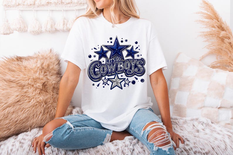 以下が含まれることがあります： 白いTシャツに、青と銀色のグラフィックで「Let's Go Cowboys」と星が描かれています。Tシャツは、白と茶色のふわふわした表面に座っている人が着ています。