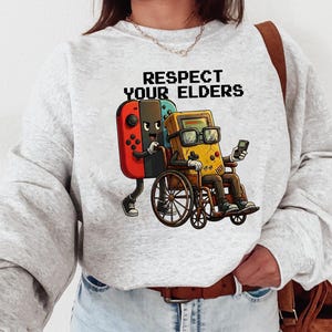 May include: A light gray sweatshirt featuring a graphic of a red and blue game controller assisting a yellow handheld game console in a wheelchair. The text "RESPECT YOUR ELDERS" is printed above the graphic in pixelated font. A fun, retro gaming theme.