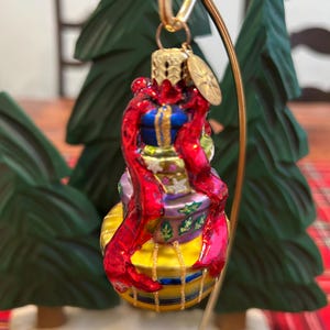 May include: A colorful Christmas ornament shaped like a stack of wrapped gifts. The ornament features a yellow base, purple and blue middle sections, and a red ribbon. It hangs from a gold-colored metal stand, with green Christmas trees in the background.