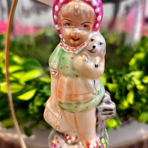 May include: A glass Christmas ornament featuring a girl in a pink and white dress holding a white dog. The ornament is decorated with green and white polka dots and has a red and white hat. The ornament is on a stand with the word "Christopher" on a tag.