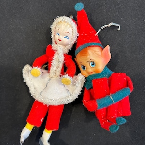May include: Two vintage Christmas ornaments: a doll in a red suit with a white fur-trimmed coat and a smiling elf in a red outfit with a green scarf. Both have painted faces and are made of felt and plastic.
