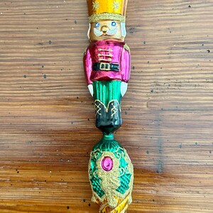 May include: A decorative Christmas ornament shaped like a nutcracker soldier. The ornament features a gold crown, a pink jacket, green pants, and a gold and green cone-shaped base. A gold star tops the ornament.