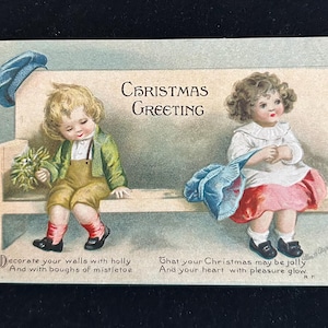 May include: Vintage Christmas greeting card featuring two children seated on a bench. The boy wears a green jacket and overalls, holding mistletoe. The girl is in a white dress with a pink skirt. Text reads "Christmas Greeting".