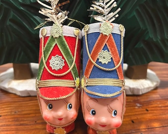 Könnte beinhalten: Zwei Vintage-Weihnachtsschmuckstücke in Form von Spielzeugsoldaten. Einer trägt einen roten Hut mit grüner Schärpe, der andere einen blauen Hut mit orangefarbener Schärpe. Beide haben goldene Verzierungen, ein lächelndes Gesicht und silberne Lametta-Akzente.