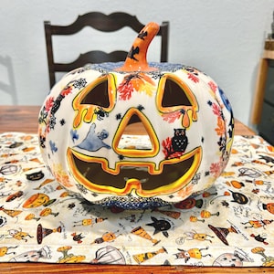 May include: A ceramic Halloween pumpkin with a carved face and decorative cutouts. The pumpkin is white with orange, yellow, and black accents, and is decorated with fall leaves, spiders, and an owl. The stem is orange with black details.