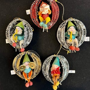 May include: A collection of vintage Christmas ornaments featuring elf-like figures. Each ornament has a silver wire frame and a small elf with a musical instrument or book. The elves wear colorful outfits, including green hats and red shoes.