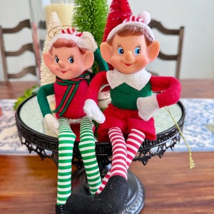 May include: Two elf dolls with painted faces, blue eyes, and pointed ears. One wears a red top with a green scarf and green-and-white striped legs. The other wears a green top with a white collar, red pants, and red-and-white striped legs.