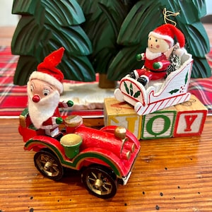 May include: Two Christmas ornaments: a red car with a Santa figure and a Santa in a sleigh. The car has gold wheels and green accents. The sleigh is white with red and green details. A wooden block spells out the word "JOY".