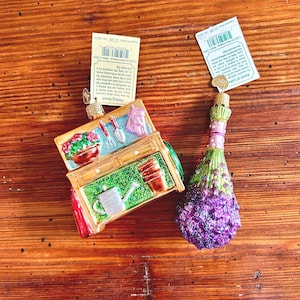 May include: Two glass Christmas ornaments with a gardening theme. One ornament is a miniature shed with a watering can, flowers, and gardening tools. The other ornament is a purple and green bunch of lavender.