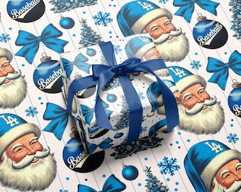 May include: A square gift box wrapped in white paper with a repeating pattern of Santa Claus wearing a blue LA baseball cap, blue bows, blue Christmas trees, and blue ornaments labeled Baseball, tied with a dark blue satin ribbon.