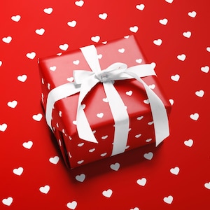 Puede incluir: Una caja de regalo roja con patrones de corazones blancos y un lazo de cinta blanco. La caja está sobre un fondo rojo salpicado de formas de corazón blancas, creando un tema romántico.
