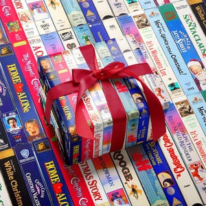 May include: A collection of vintage VHS tapes featuring classic holiday films arranged in a grid pattern, with a central stack wrapped in a bright red satin ribbon and bow. The spines display titles like Home Alone, Elf, and White Christmas.