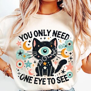 Op de afbeelding: Een wit T-shirt met een zwarte kat illustratie en de tekst "You only need one eye to see". De kat heeft één groot blauw oog en is omringd door sterren, manen en andere hemellichamen.
