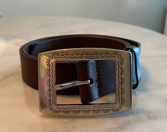 May include: A brown leather belt with a silver buckle featuring a decorative design. The buckle is rectangular with a detailed border.