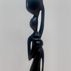 May include: A tall, dark wooden sculpture of a woman carrying a vessel on her head. The figure has a slender form with flowing lines, and the sculpture is mounted on a square base. The artwork is a decorative piece.