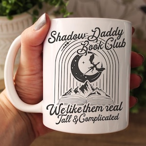May include: White ceramic mug with a black design that says "Shadow Daddy Book Club" with a crescent moon, a dragon, and mountains. The text below the design says "We like them real tall & complicated."