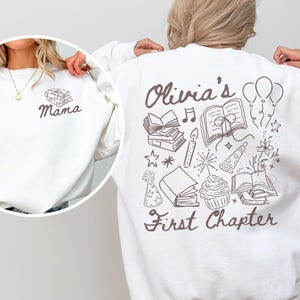 May include: A white long-sleeve sweatshirt featuring dark brown line art. The back displays the text "Olivia's First Chapter" surrounded by illustrations of books, balloons, a cupcake, and stars. The front chest shows a small stack of books and "Mama".
