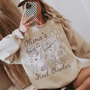 May include: A beige crewneck sweatshirt featuring a dark brown graphic print with the text 'Olivia's First Chapter'. The design includes illustrations of open books, a cupcake, balloons, a party hat, a candle, and stars, creating a celebratory theme.