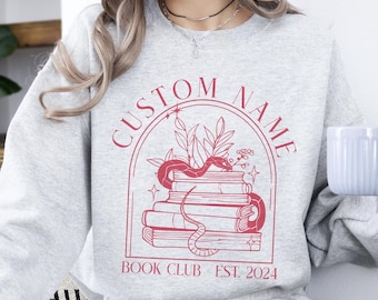 May include: Gray sweatshirt with a red graphic of a snake curled around a stack of books. The graphic also includes a floral arch and the text "CUSTOM NAME BOOK CLUB EST. 2024".