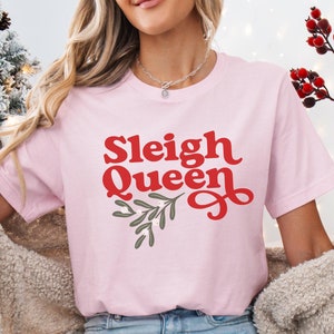 May include: A pink t-shirt with the text "Sleigh Queen" in red letters with a green holly sprig design. The shirt is being worn by a person with long blonde hair.