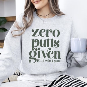 May include: A grey sweatshirt with the text "zero putts given jk... it was 4 putts" in green lettering. The person wearing the sweatshirt is holding a light blue mug.
