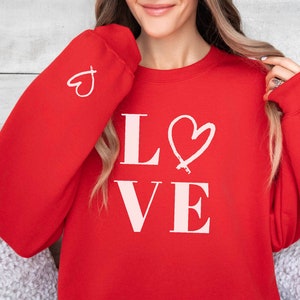 Peut inclure: Sweat-shirt rouge avec un graphisme "LOVE" rose et un cœur rose sur chaque manche.