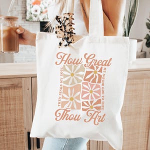 May include: Cream-colored tote bag with the text "How Great Thou Art" in a stylized font. The design includes floral patterns in shades of peach, pink, and cream. The bag has a simple, minimalist design with long handles.