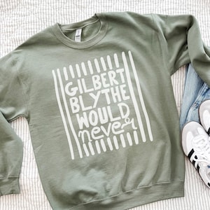 May include: An olive green crewneck sweatshirt featuring white screen-printed text that reads "GILBERT BLYTHE WOULD NEVER" framed by vertical lines. The sweatshirt is paired with blue denim jeans and white trainers with black stripes.
