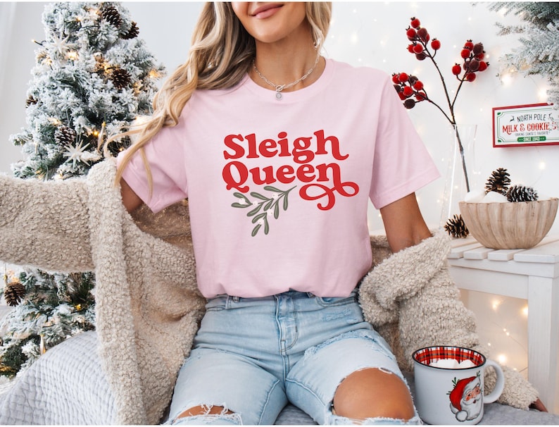 May include: A pink t-shirt with the text "Sleigh Queen" in red letters with a green holly sprig design. The shirt is being worn by a person with long blonde hair.